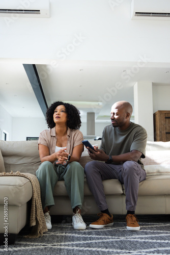 Obraz na płótnie African American couple sitting on light gray sofa in open plan living area hold