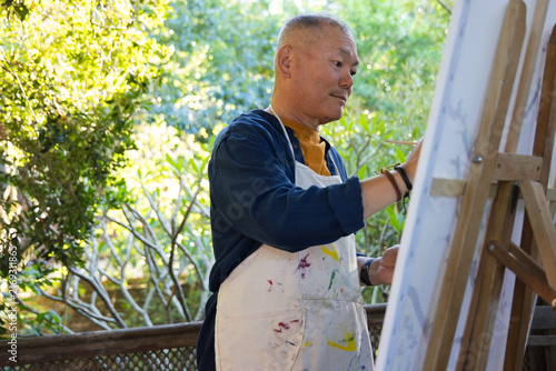 Bild auf Leinwand Senior Asian man holding brush standing in garden painting canvas at easel weari