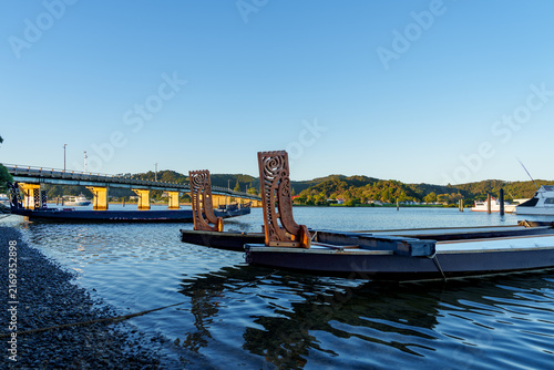 Decorated prow or bow piece of Maori waka on beach with Waitangi one way Bridge in silhouette back-lit by morning sun in background