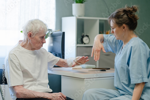 Hand rehabilitation therapy for elderly man with broken arm and cast in hospital. Physical therapist guiding senior patient through recovery exercise with gentle focused dedication indoors