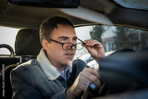 Male driver adjusting his glasses and looking ahead with a focused expression while driving. Correcting vision, paying attention to the road, and safe driving habits.
