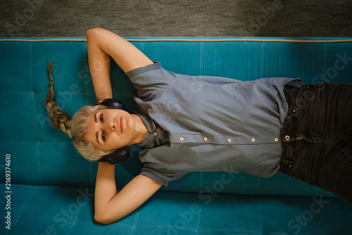 Woman lies on blue couch with hands behind head. She wears headphones and enjoy music. Natural light fills space, creating relaxed atmosphere for leisure and rest.