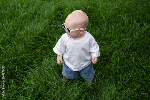A charming boy wearing sunglasses is learning to walk on the green grass. The little one is walking barefoot on the lawn.