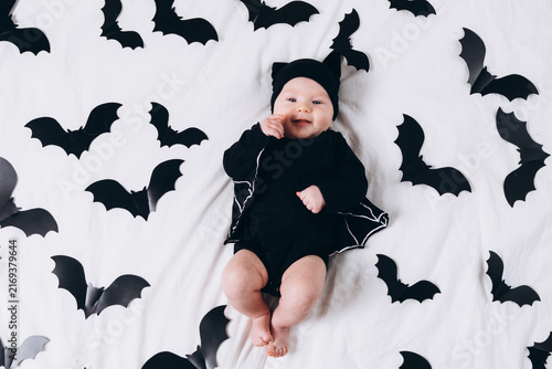 A cute baby in a bat costume lies on a white background featuring a bat pattern. A Halloween costume for a baby.