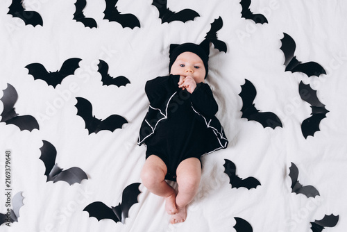 A cute baby in a bat costume lies on a white background featuring a bat pattern. A Halloween costume for a baby.