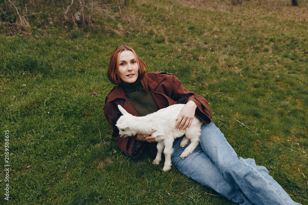 Naklejka premium Relaxed woman lies on grass with a playful goat kid beside her, capturing countryside vibes and gentle companionship. Casual fashion, cozy mood, and natural lighting convey a wholesome lifestyle