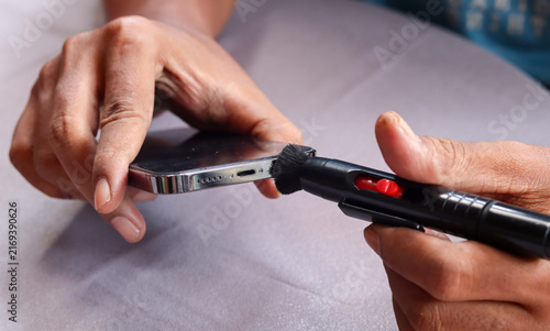 Asian man's hand cleaning a smartphone speaker grille with a small cleaning brush, also brushing around the charging port to remove dust, lint, and debris for better device maintenance and care.