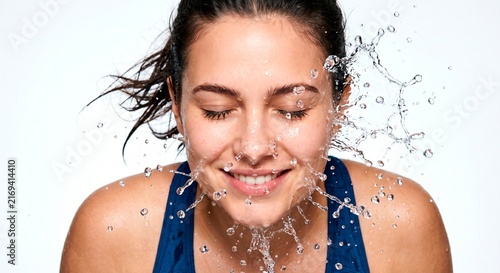Happy young woman splashing fresh clear water on her face with eyes closed, dynamic flying droplets on a white background, hydration and skincare concept.
