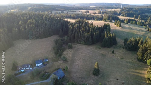 Smooth aerial view over lush green countryside with open fields, dense trees, winding rural roads and peaceful natural scenery in bright daylight.