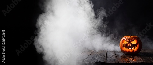 Terrifying carved halloween pumpkin with menacing grin and glowing eyes surrounded by thick white smoke on wooden surface
