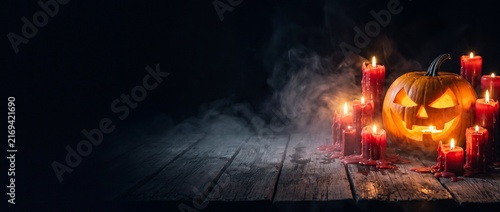 Fierce glowing jack lantern surrounded by melting red candles on wet dark wooden planks under heavy fog for spooky halloween wallpaper