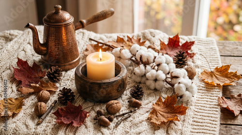 Cozy autumn scene with a burning candle, copper pot, cotton bolls, fallen leaves, and pinecones on a knit blanket, symbolizing warmth and comfort