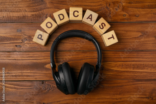 Black wireless headphones surrounded by wooden blocks spelling PODCAST on a dark wooden background. Top view flat lay.
