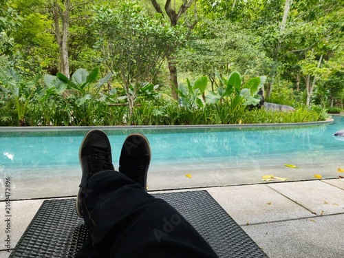 A first-person point-of-view (POV) shot of a person relaxing on a poolside lounger with feet crossed, overlooking a clear blue swimming pool framed by lush tropical foliage and dense green trees 