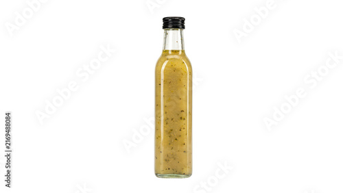 A tall, slender clear glass bottle filled with yellow salad dressing or vinaigrette, containing visible herbs and spices, with a black cap, isolated