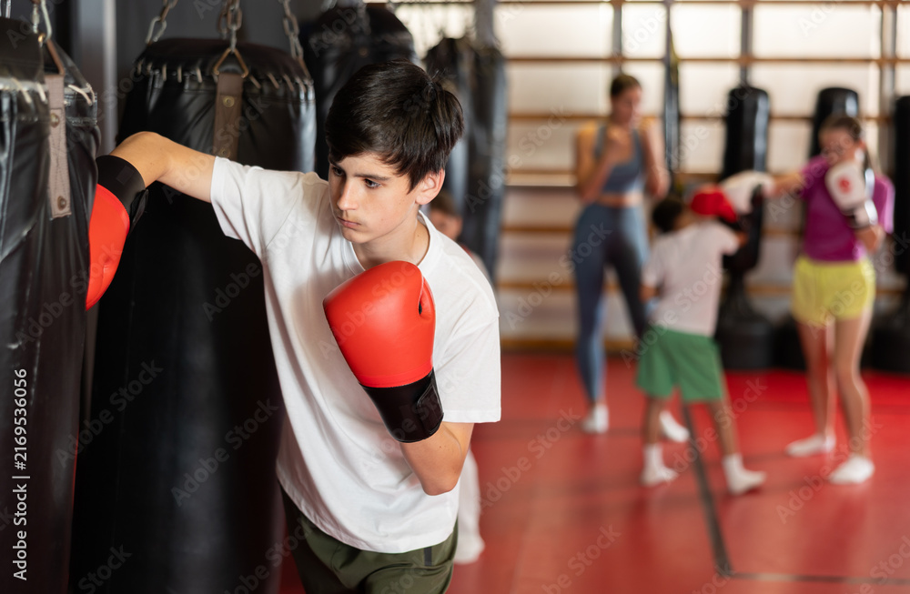 Fototapeta premium Boy in red boxing gloves stands near a punching bag. Child in boxing training learns punches for self-defense