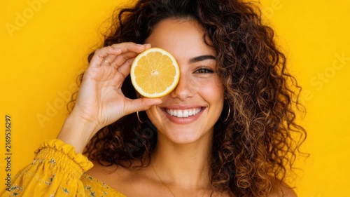 Happy young woman with curly hair holding lemon slice over eye smiling against bright yellow background