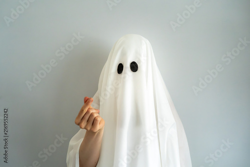 Playful bedsheet ghost making a Korean finger heart gesture, combining Halloween, love and affection with dynamic hand movement against a minimalist background.