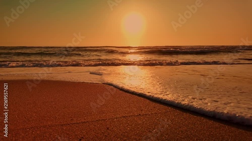 A serene sunset casts a warm orange glow over the ocean as gentle waves wash onto a wet sandy shore. The peaceful coastal scene evokes a sense of calm and relaxation.