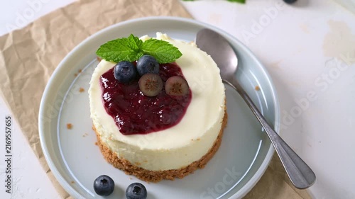 A delicious mini cheesecake topped with rich berry compote, fresh blueberries, and mint leaves sits on a light blue plate.