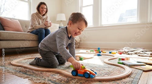 Little boy playing with toy car on wooden train track while mother watches from sofa