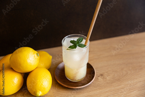 Citronnade glacée servie dans un verre transparent avec une paille et des feuilles de menthe, table en bois, fond brun, citrons jaunes posés à gauche du verre, vue en plongée, sensation de fraîcheur 