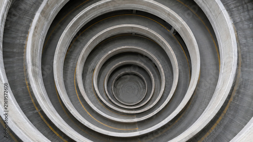 Aerial view looking straight down into a concrete spiral parking garage, its concentric circular ramps and yellow lane lines forming a hypnotic vortex descending toward the dark center.