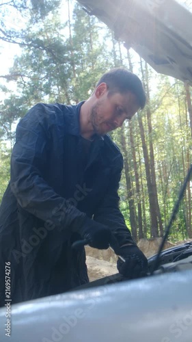 Focused mechanic in uniform and gloves using socket wrench to repair car engine with hood up, standing on road in sunny forest, demonstrating professional vehicle maintenance and repair. Vertical shot