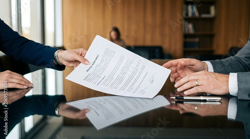 Business professionals exchanging a signed legal agreement across a reflective desk surface inside an office meeting room while collaborating on important financial documents and corporate