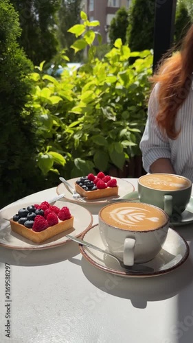 Close-up outdoor cappuccino with latte art and berry tarts on ceramic tableware, surrounded by green foliage and sunlight, cozy summer café atmosphere, lifestyle food photography.