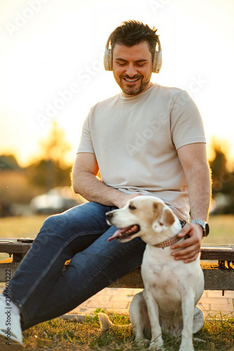 Man wearing headphones and petting his dog while relaxing on a bench at sunset