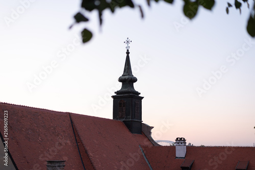 Quadro su tela Historic Petrovaradin church spire rises above red-tiled roof against soft gradient sky at twilight