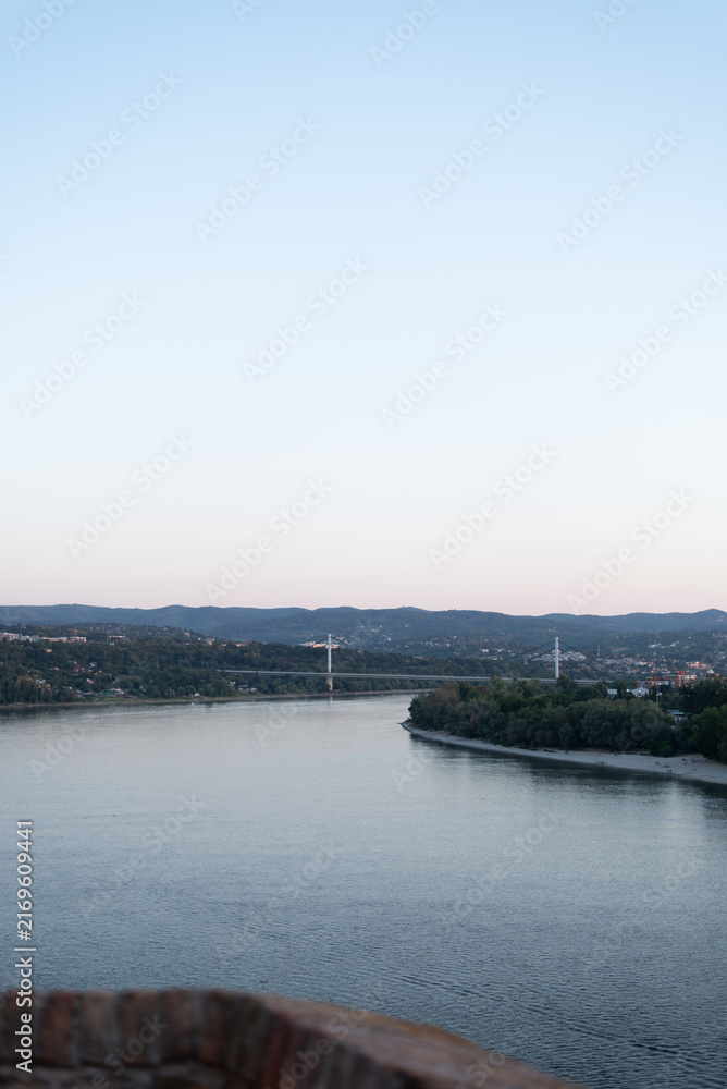 Obraz premium Wide Danube river flows through hilly landscape with bridge spanning across in the distance. Novi Sad, Serbia
