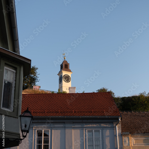 Historic white clock tower of Petrovaradin Fortress rises behind the red tiled roof of building. Novi Sad, Serbia