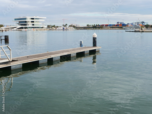 Empty wooden floating dock in Marina of Valencia with modern pavilion building in distance. Beautiful scene for Mediterranean yachting lifestyle blogs, luxury travel guides or coastal tourism banner