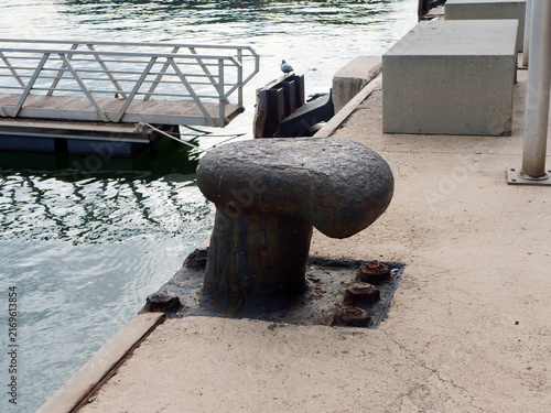 Old rusty cast iron mooring bollard on concrete pier with floating dock in background. Useful element for maritime transport industry article, port logistics website or coastal travel blog banner