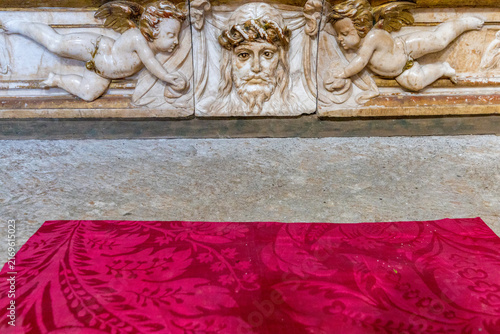An intricate marble relief featuring cherubs and a sacred face lies above a red patterned cloth
