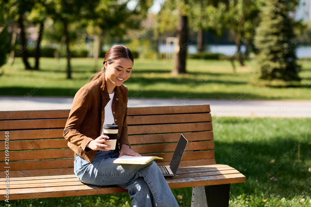 Fototapeta premium A young beautiful Asian woman leisurely sips coffee, absorbed in her work at a sunny park.