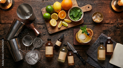 Top down view of professional mixology bar setup with cocktail shaker jigger strainer ice fresh citrus fruits mint bitters bottles and notebook on dark wooden counter
