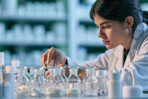 A focused female scientist in a white lab coat carefully handles glass bottles filled with white pills in a brightly lit professional pharmacy laboratory setting
