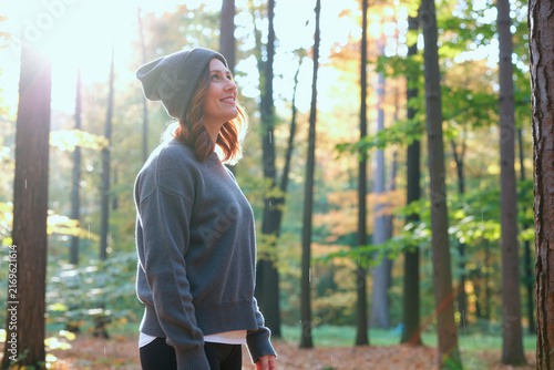 Young adult Caucasian woman standing in forest, looking upward and smiling, expressing mindfulness and outdoor wellness. Ideal for mental health, self care, nature retreat marketing