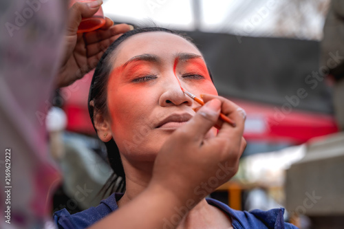Unrecognizable makeup artist applying vibrant red paint in traditional opera style to  female actress backstage while preparing for a classic Chinese opera stage performance