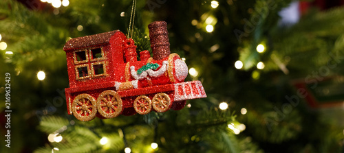 Fotografie Red glitter toy train ornament hanging on a lit christmas tree with warm bokeh, panoramic crop ideal for holiday banners, party invitations and seasonal web headers