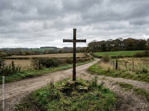 Wooden cross stands at a rural crossroads under a cloudy sky.