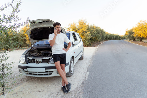 Fotografie Young man standing by his broken down car with open hood, calling for roadside a
