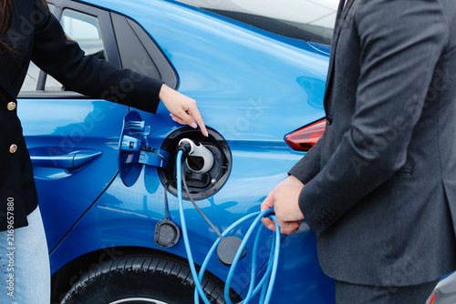 Sales manager showing how to charge electric car to a young client woman, plugging wire into the car socket at the car dealership outdoor