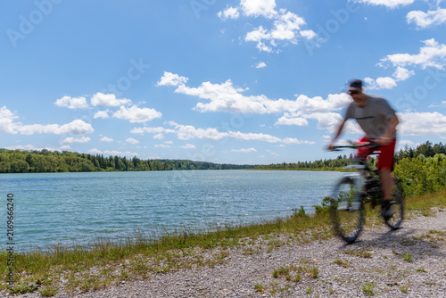 Motion blurred cyclist riding mountain bike along scenic lake shore under blue sunny sky, active lifestyle and outdoor sports concept