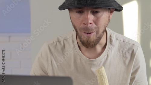 man eating banana in indoor setting with laptop and cloth shirt, neutral mood, possibly in office or studio, frontal view, beige and black colors, commercial food and beverage context.