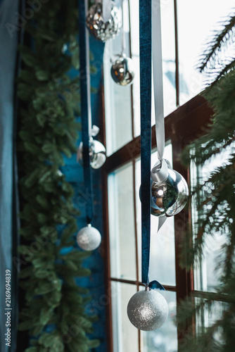Silver glass Christmas baubles hanging on ribbons near window with spruce garland.