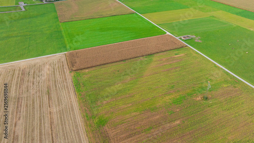 High angle aerial shot displays scenic rural landscape of Lind im Drautal village. Winding Drava river, agricultural fields, main road, and railway line cross lush green valley surrounded by forested 